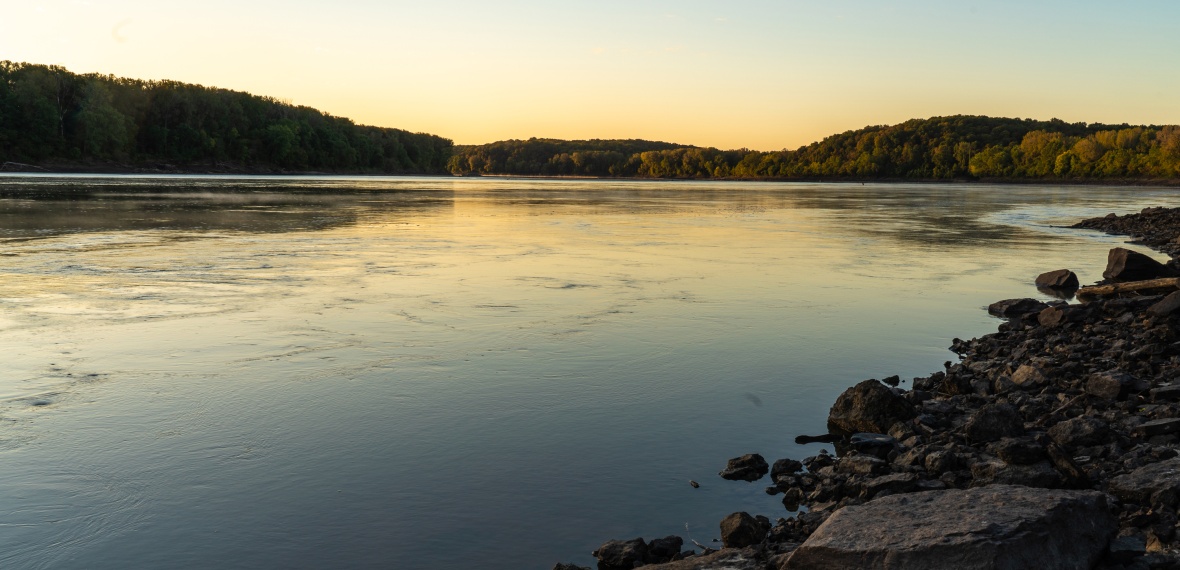 Water flows down the Missouri River. The rocky bank is in the foreground and the sun is setting behind trees in the background.