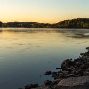 Water flows down the Missouri River. The rocky bank is in the foreground and the sun is setting behind trees in the background.