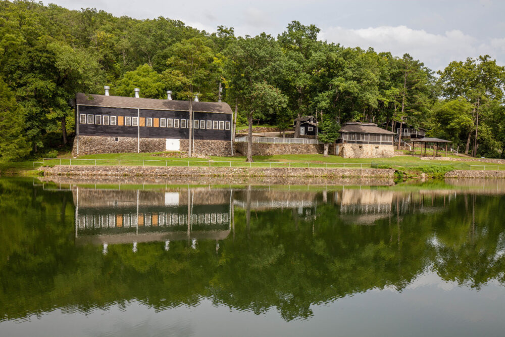 In the foreground is a calm lake. Across the lake, in the background, are a series of buildings with fences and walkways between them. Trees surround the buildings.