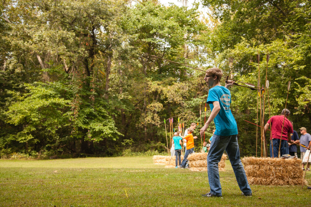 A teenage boy throwing an atl atl, which is a 4-5 ft. spear that has a 12-18 inch. throwing handle. Groups of children and instructors are in the background among hay bales and spears stuck vertically into the bales.