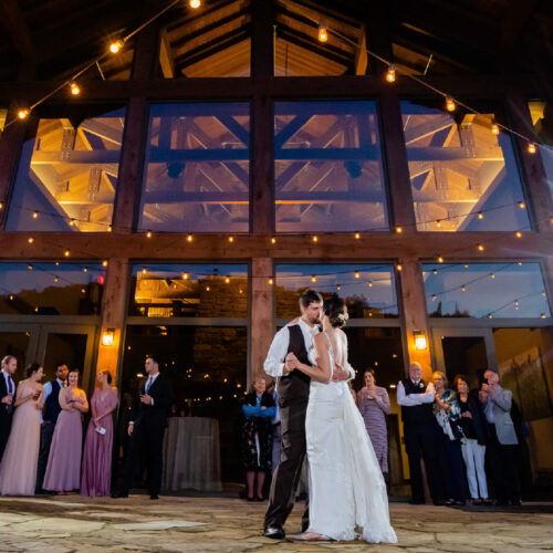 A newly married couple at their wedding lean in for a kiss, surrounded by a group of family and friends. In the background is a large lodge.