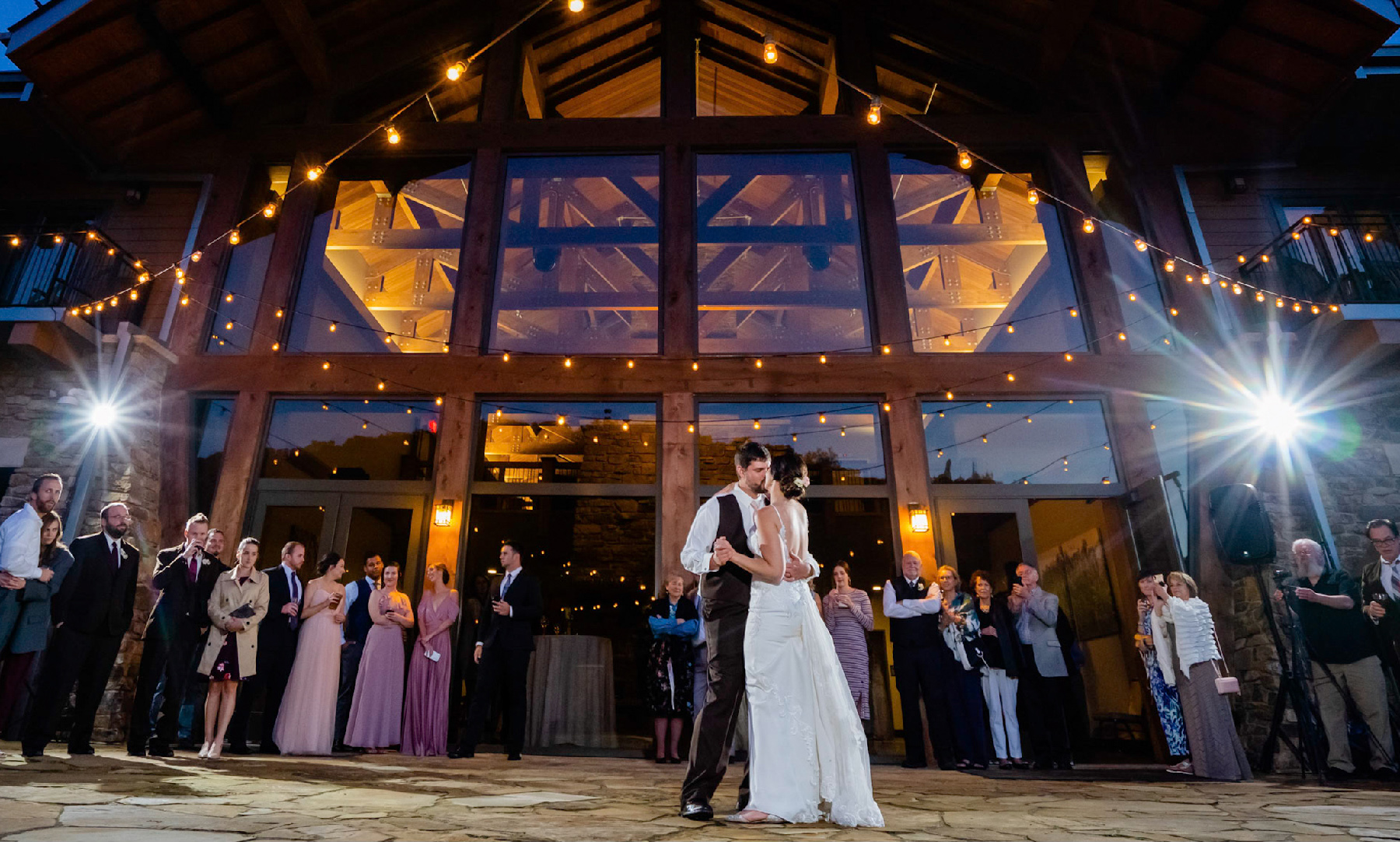 A newly married couple at their wedding lean in for a kiss, surrounded by a group of family and friends. In the background is a large lodge.