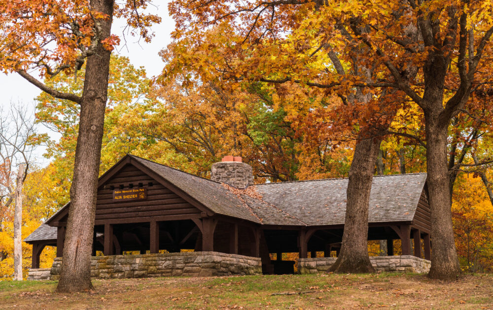 A large open-air shelter constructed of brown timbers. The base is made of limestone bricks. The shelter is nestled among tall oak trees showcasing their fall foliage.