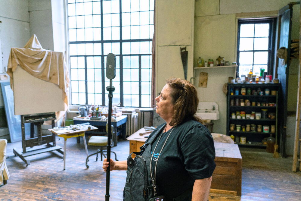 A woman stands in Thomas Hart Benton's studio holding a 360-degree camera attached to a pole above her head.