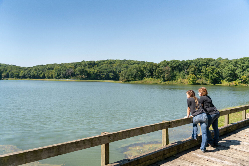 Two people stand on a wooden overlook that looks out over the lake.