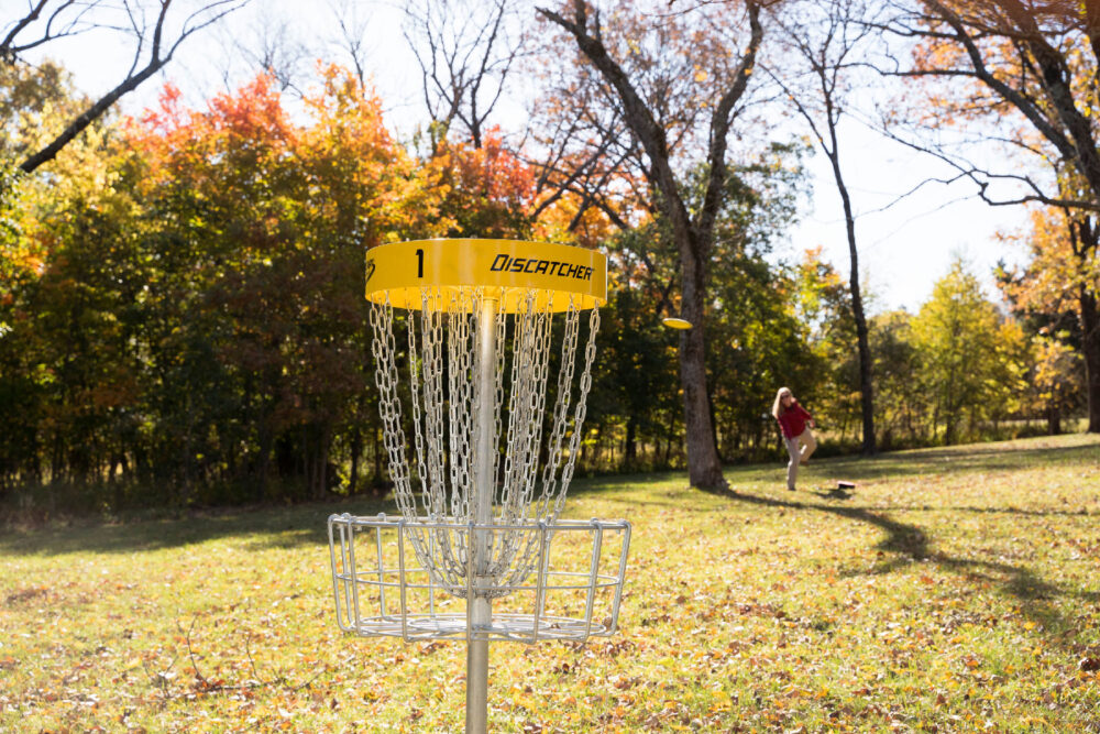 An image of the basket. Metal chains hang down from a pole that sticks out the top of the basket. In the background there is a person throwing a disc. The disk has already left their hand and if flying through the air toward the basket.