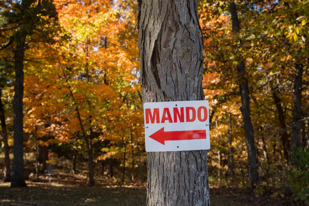 An image of a sign attached to a tree. In large letters, it says MANDO. Underneath the sign text is an arrow pointing to the left.