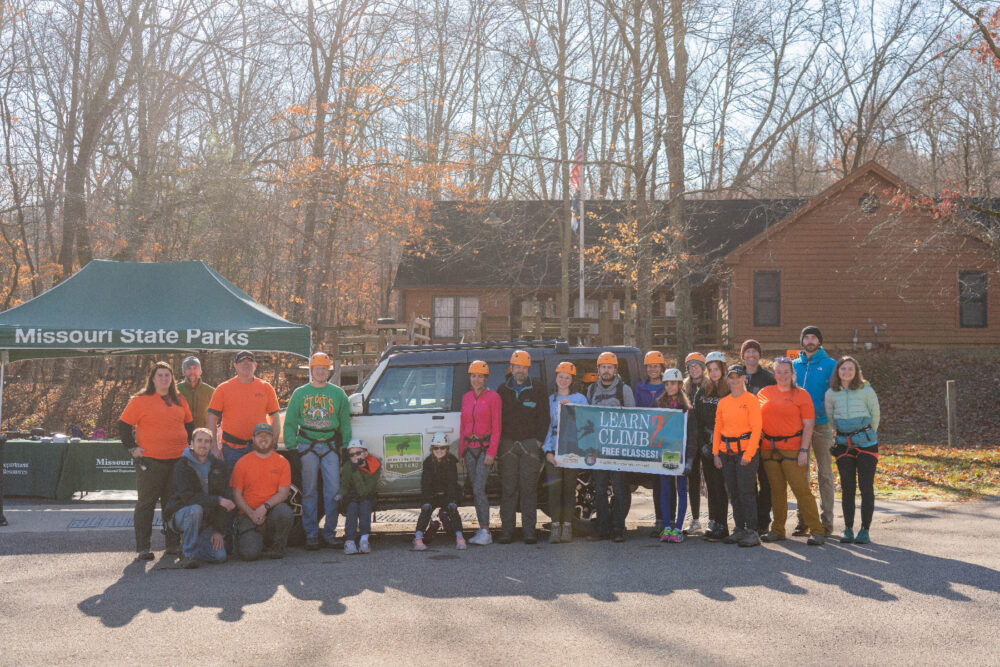 A group of 21 people stand in front of a Ford Bronco SUV holding a Learn2 Climb sign. The SUV has a Bronco Wild Fund logo. In the background is a pop-up shade tent with Missouri State Parks insignia. In the extreme background is the visitor center building with a flagpole.