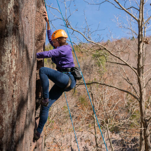 A woman wearing a helmet and a harness with support ropes climbing up a rock face.