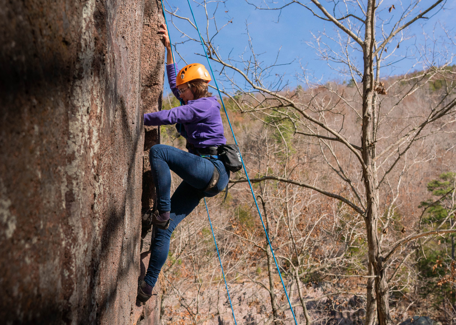 A woman wearing a helmet and a harness with support ropes climbing up a rock face.