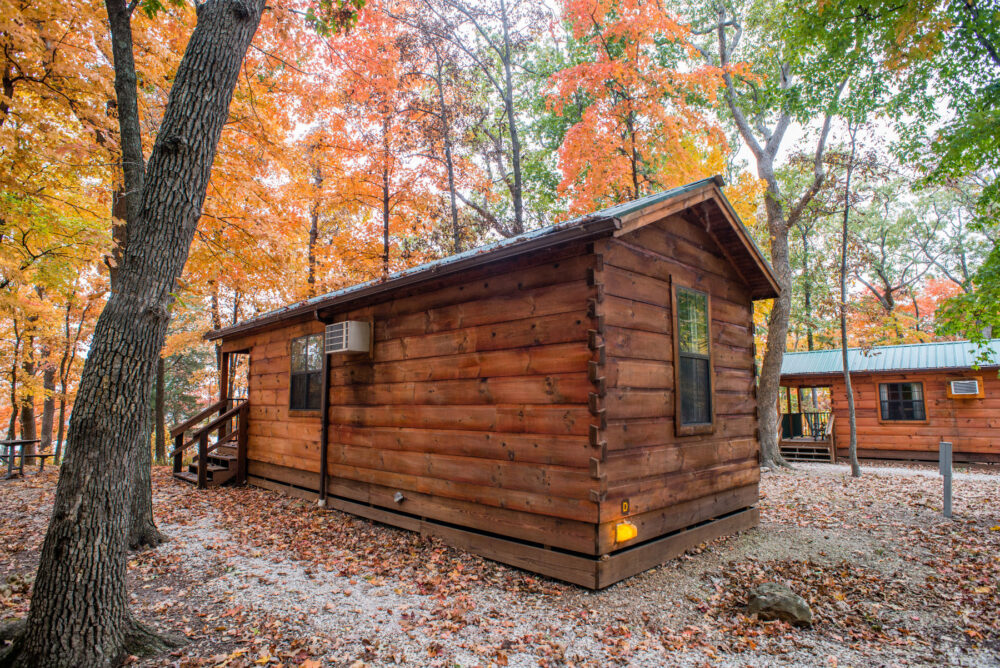 A log cabin is in the center of the frame, shaded by trees. In the background to the right is another cabin. On the extreme left edge of the frame is a picnic table.