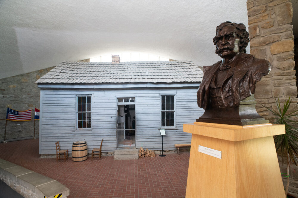 A small house with two windows and a door. The house has been moved to the interior of a museum. In the foreground is a bronze bust sculpture of Mark Twain sitting on a wooden pedestal.