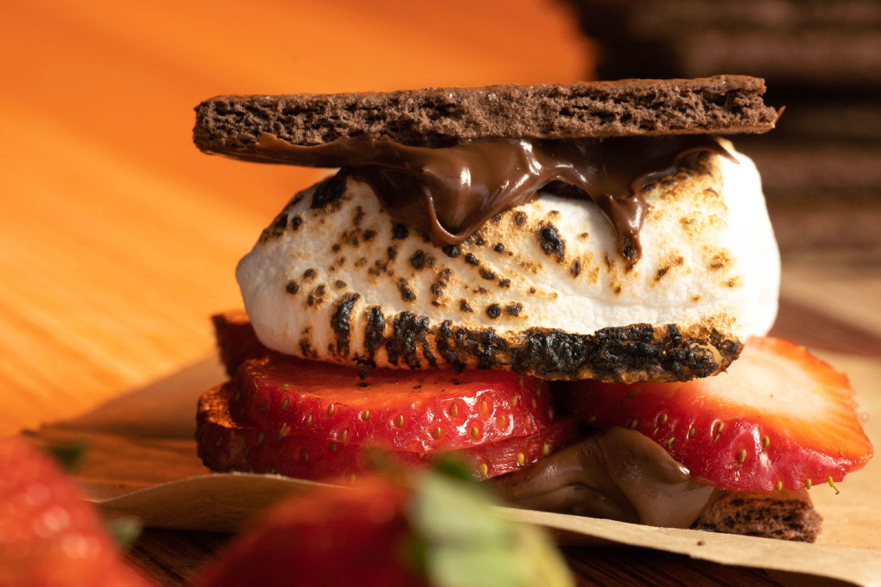 A smore. The layers of the treat can be seen. From the top, chocolate graham cracker, hazelnut spread, roasted marshmallow, sliced strawberries, hazelnut spread and a chocolate graham cracker on the bottom.