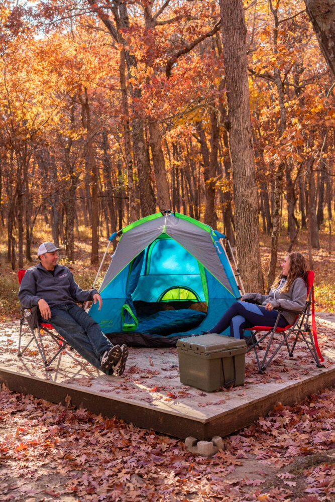 Two people sit in chairs by their tent. The tent and chairs are on a wooden platform. The area is wooded and the trees are transitioning to fall foliage.