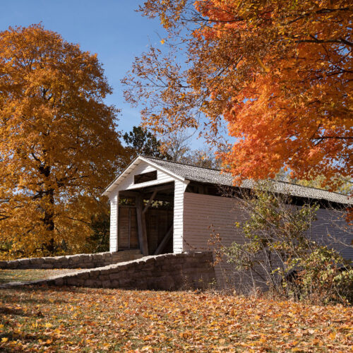 The entrance to a wooden covered bridge is in the center of the frame. To the right and left of the entrance to the bridge are large trees transitioning to fall foliage.