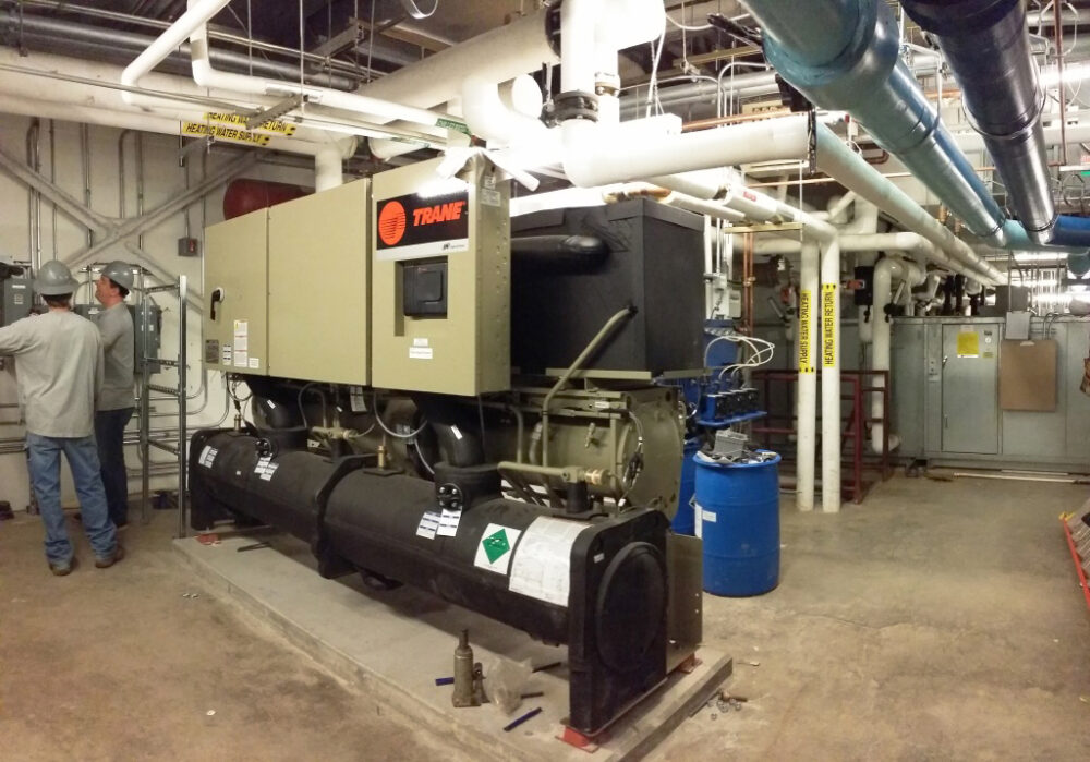 A large TRANE HVAC unit in a mechanical room of a hospital building. Two maintenance workers are on the left edge of the frame examining an electrical panel.