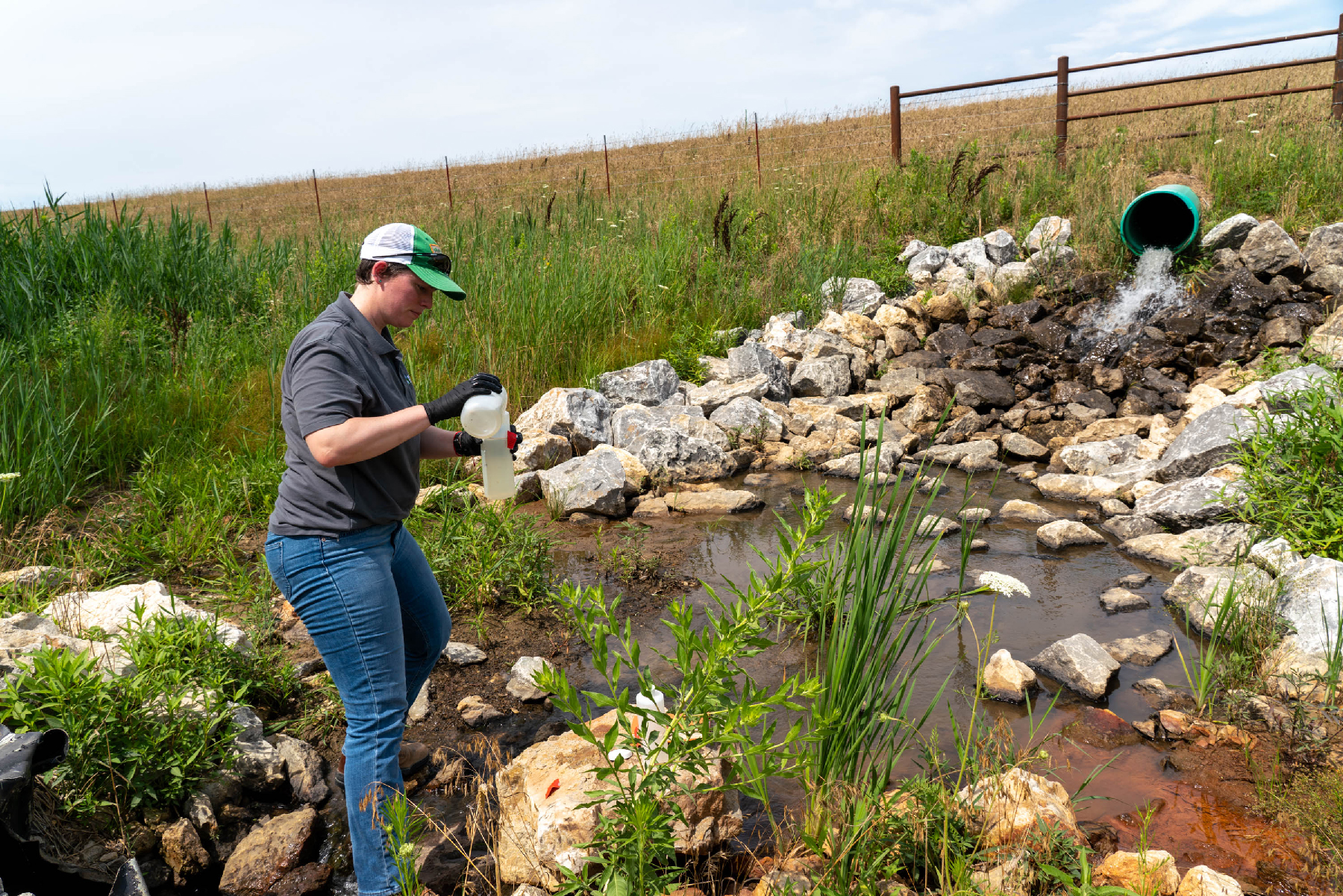 A woman stands below a culvert that is empting water onto the ground. Large limestone rocks are below the culvert to prevent erosion. The Woman is taking water samples and is holding two plastic water collection bottles.