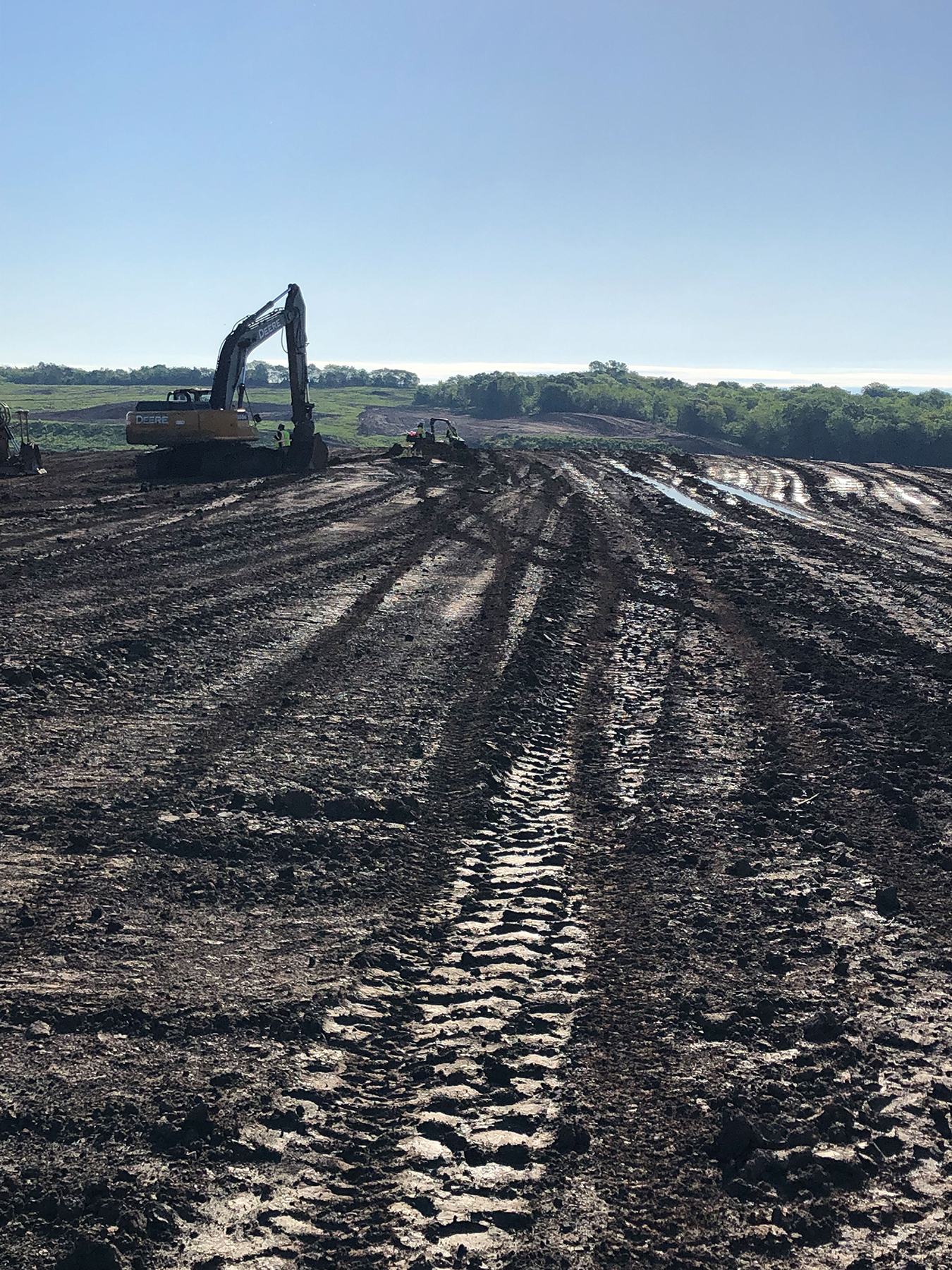 An open area that is the construction site for a new reservoir, with a muddy strip in the middle. The muddy section is crisscrossed with tracks from heavy equipment treads. In the distance is a large backhoe that has been spreading dirt.