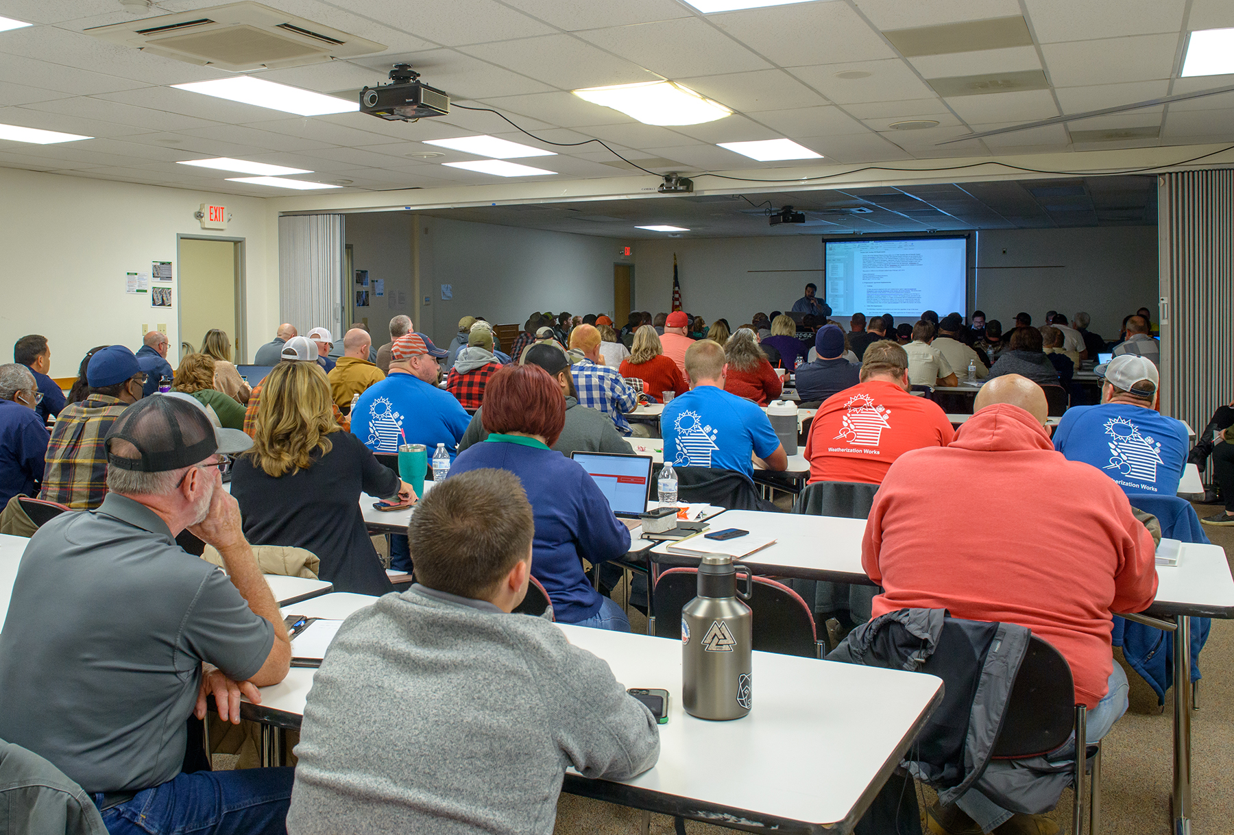 A large conference room filled with people sitting at tables. At the far end of the room is a projection screen and an instructor standing at a podium.