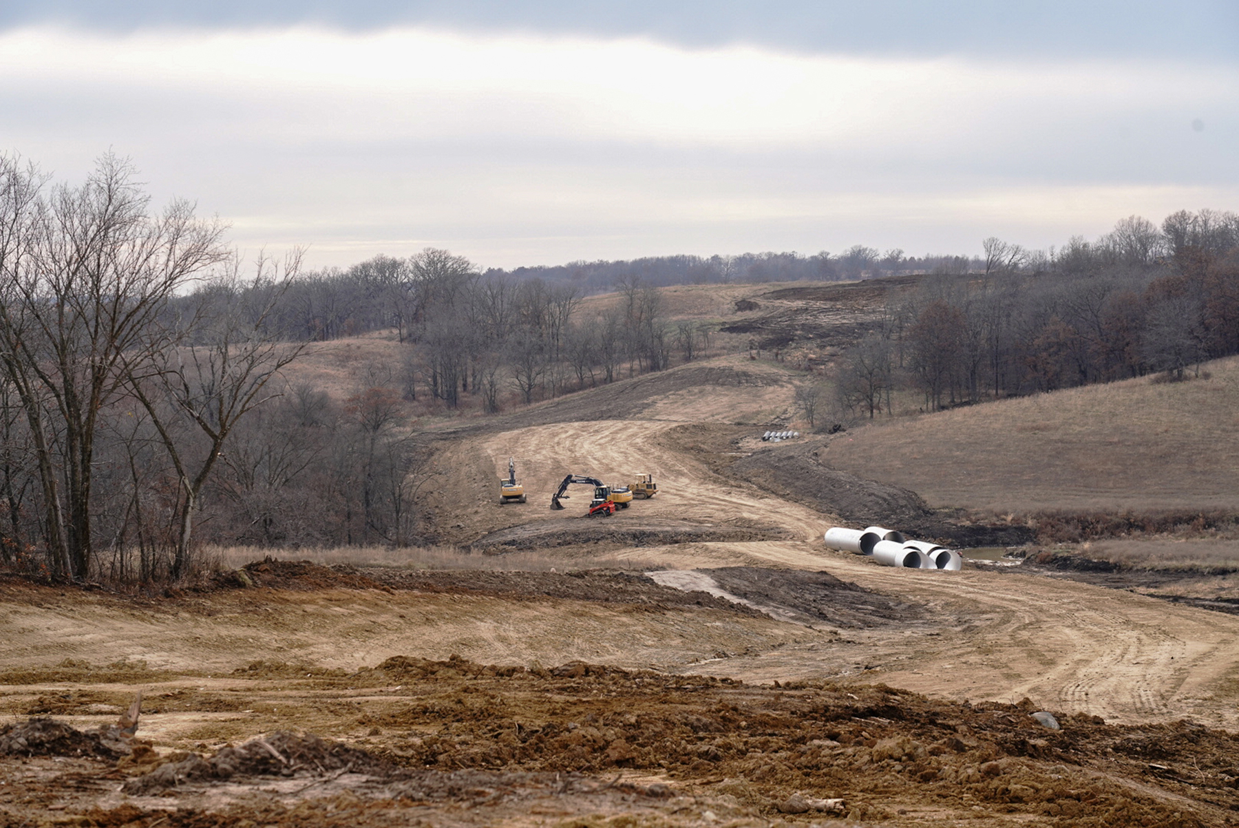 A landscape image of a valley nestled among hilly terrain. The earth is disturbed and heavy equipment tracks are visible. Two backhoes are in view, as well as a few concrete pipes that are staged for placement.