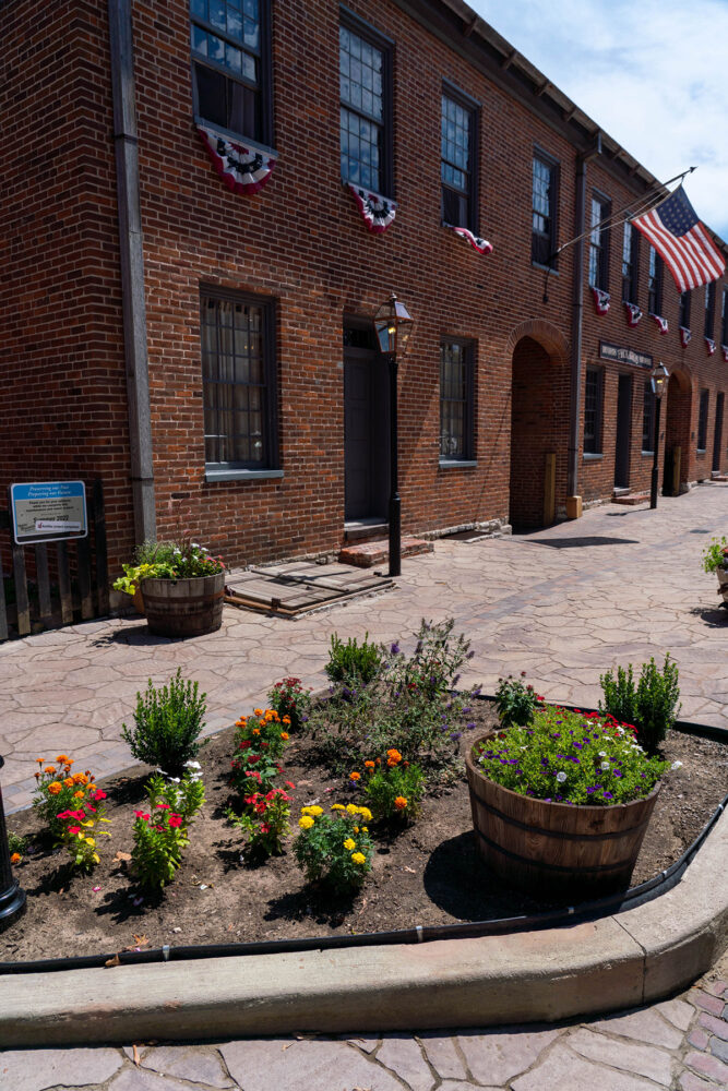 A brick building in the background with a newly-landscaped flower bed in the foreground. The sidewalks are concrete but resemble cobblestone.