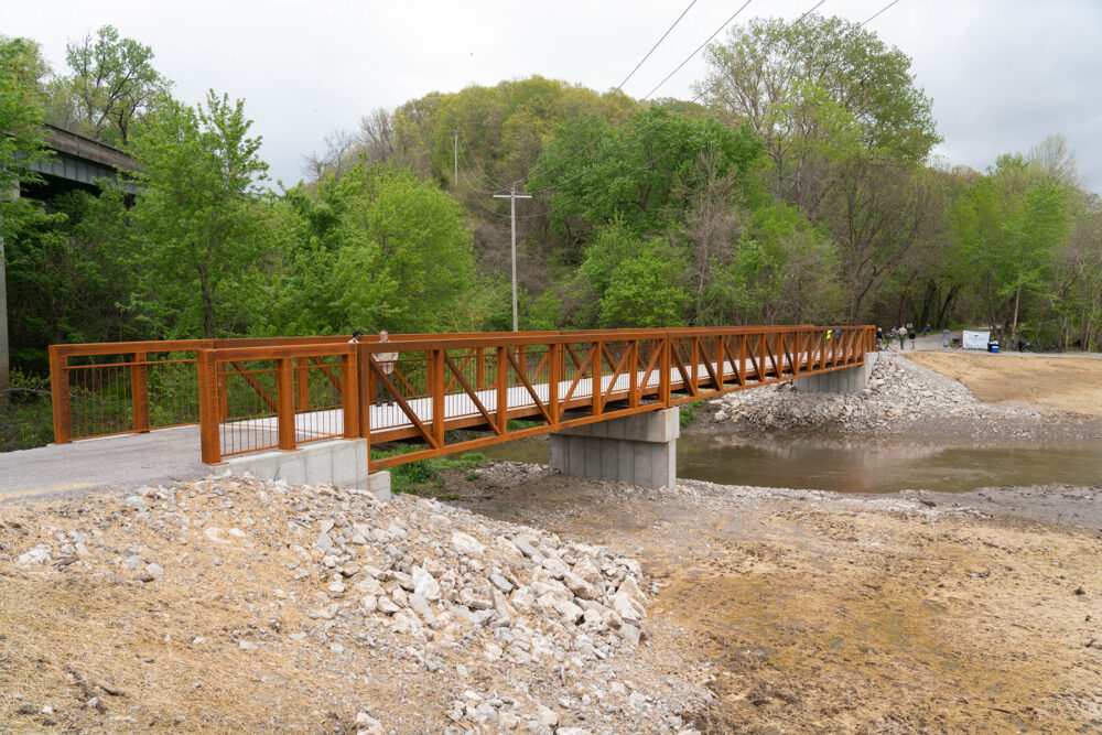 A concrete walkway extends over a creek. The railings are made from iron that has oxidized.