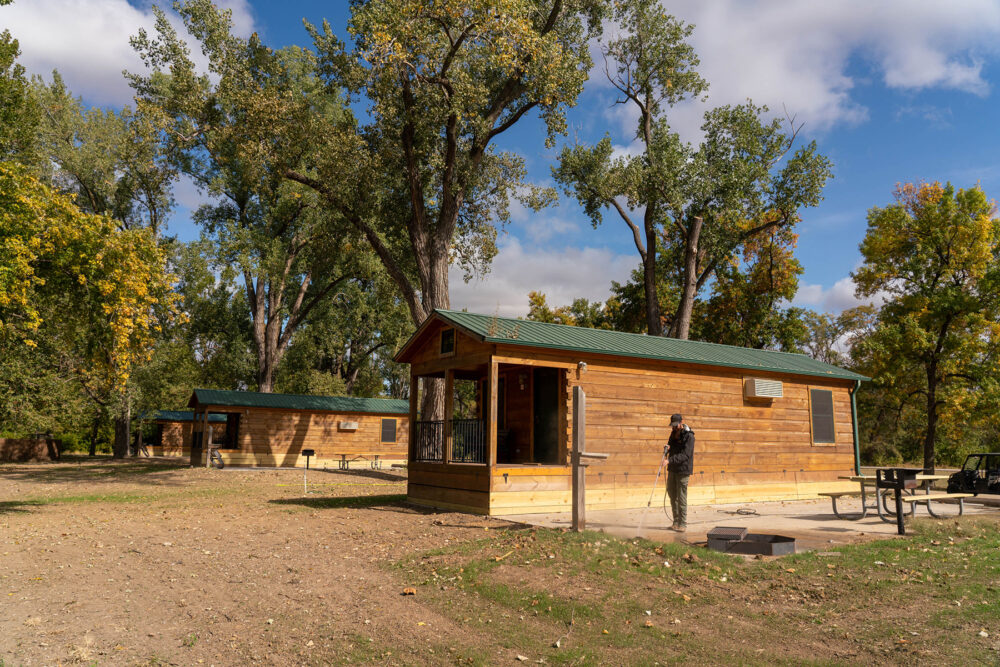 A small wooden cabin with a man standing in front of it. There is a picnic table and BBQ grill on the left edge of the frame.