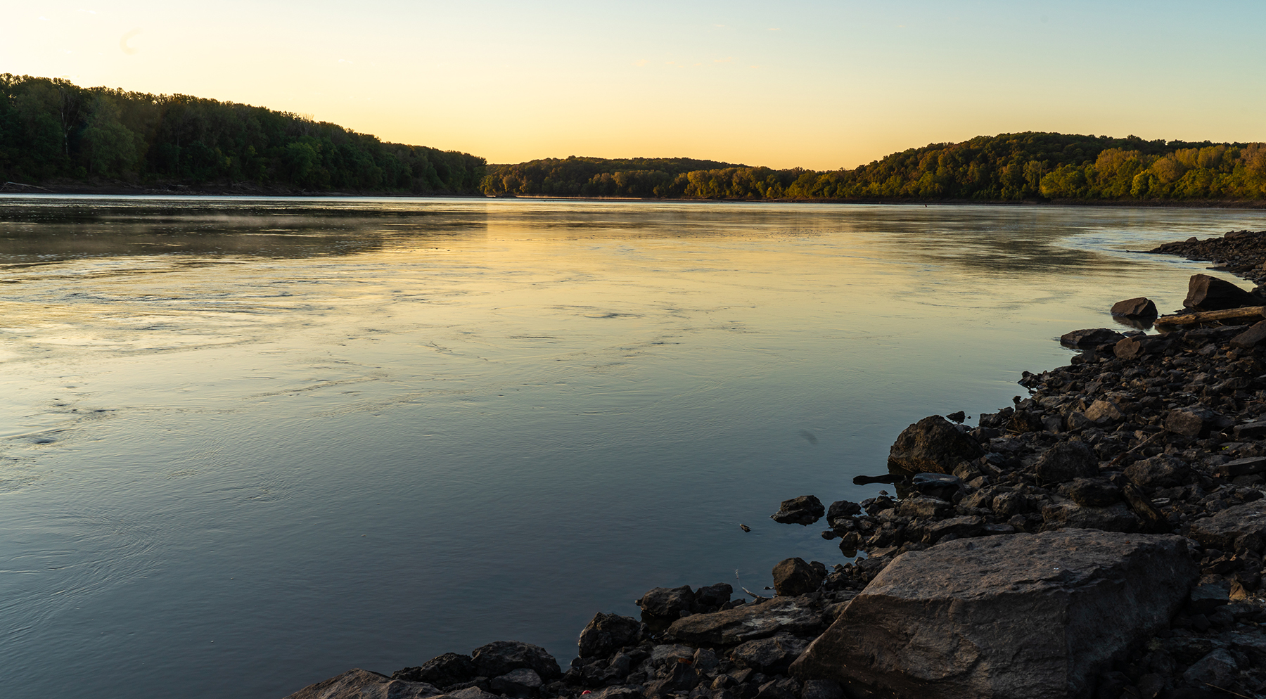 A landscape image of a river a sunset. In the foreground is the riverbank covered in large pieces of rock. In the background is a hilly treeline on the opposite bank of the river. The setting sun is reflecting off of the water in the center of the image.