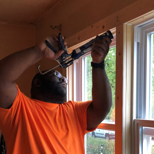 A man standing in a window inside a house. He's holding a caulking gun above his head and applying caulk around a window frame.
