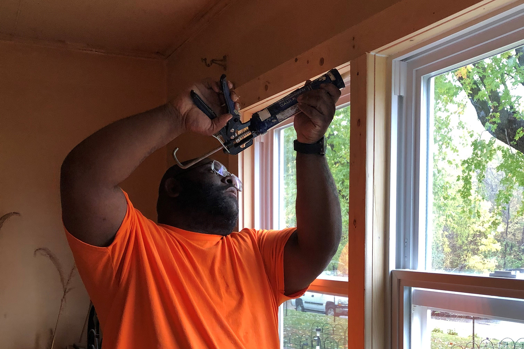 A man standing in a window inside a house. He's holding a caulking gun above his head and applying caulk around a window frame.