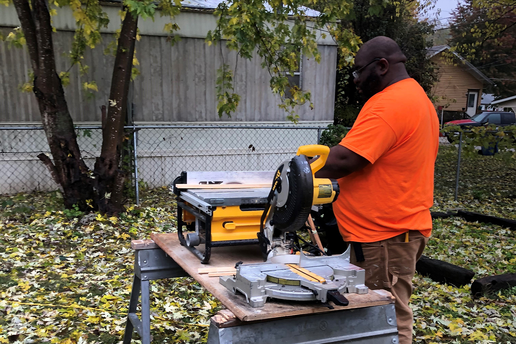 A man stands in front of a portable table saw set up outside of a house. He is feeding a piece of wooden window trim through the saw.