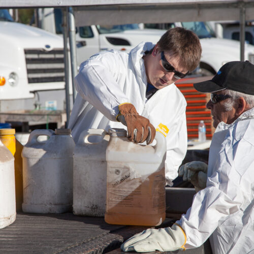 Two men wearing white protective jumpsuits, gloves and eye protection are handling containers of pesticide as they load them onto the bed of a pickup truck.