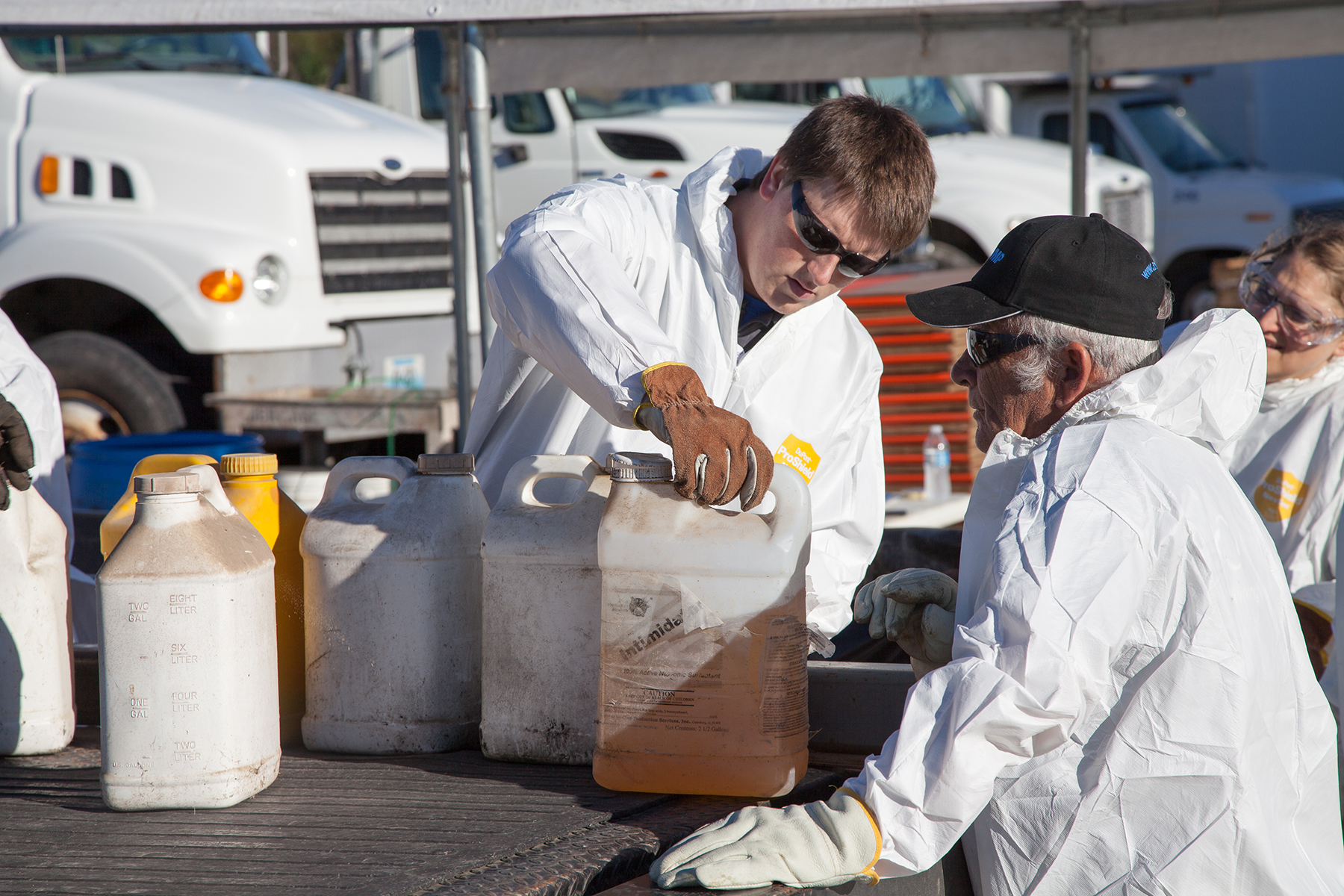 Two men wearing white protective jumpsuits, gloves and eye protection are handling containers of pesticide as they load them onto the bed of a pickup truck.