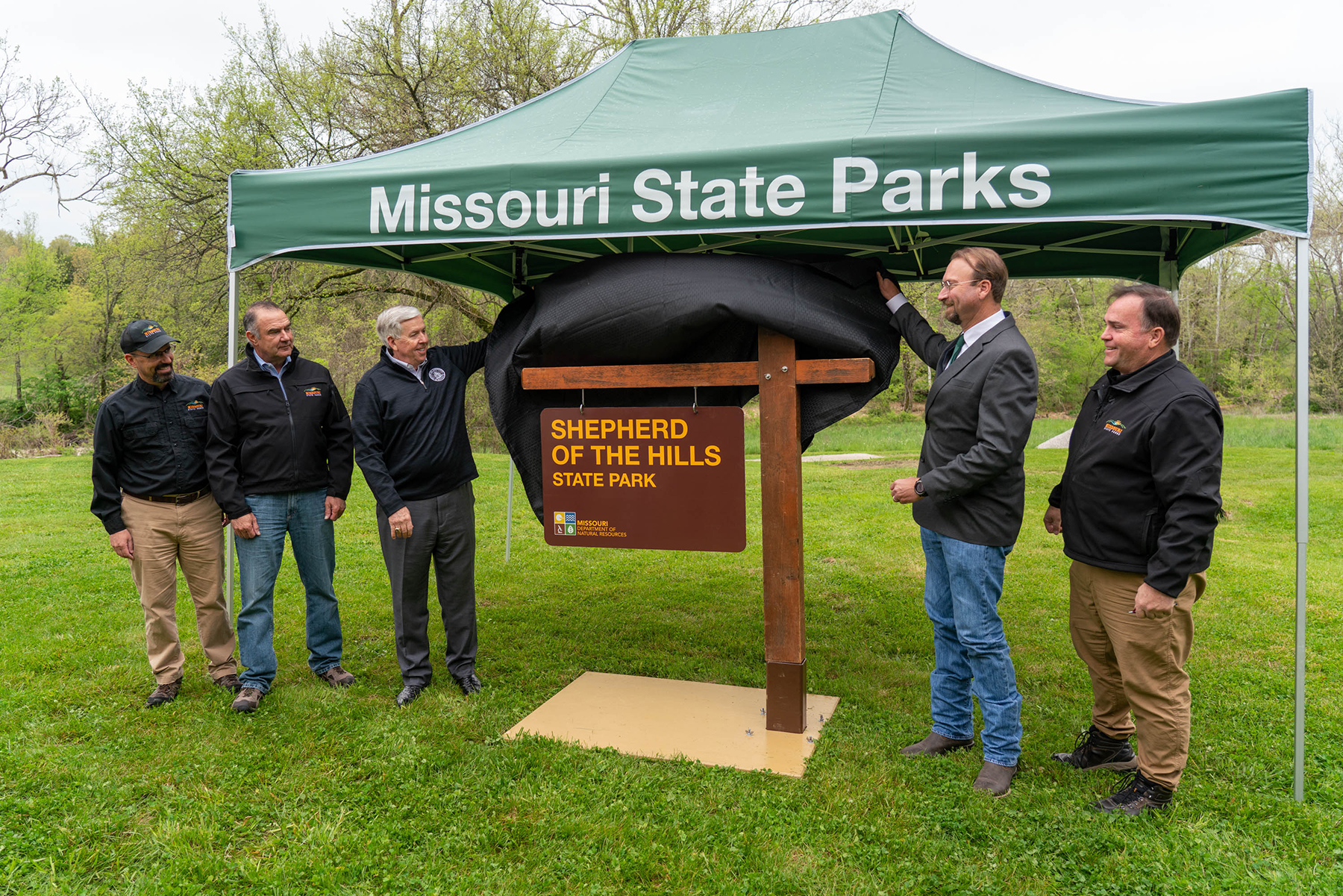 Five men stand under a popup tent. There is a cantilever-style sign in between them. The men are moving a piece of fabric that was covering the sign out of the way.