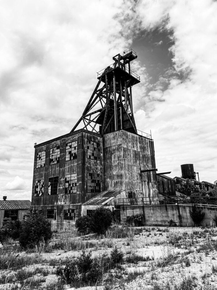 A large building covered with rusty sheet metal towers over scrub brush. Many of the windows panes on the side of the building are broken.