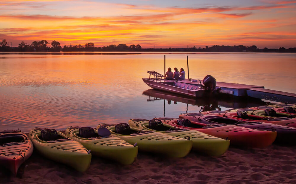 A sunset view overlooking the water. A row of kayaks line the foreground. In the center, right of the image, there is a dock with a small boat. Three people set on the end of the dock.