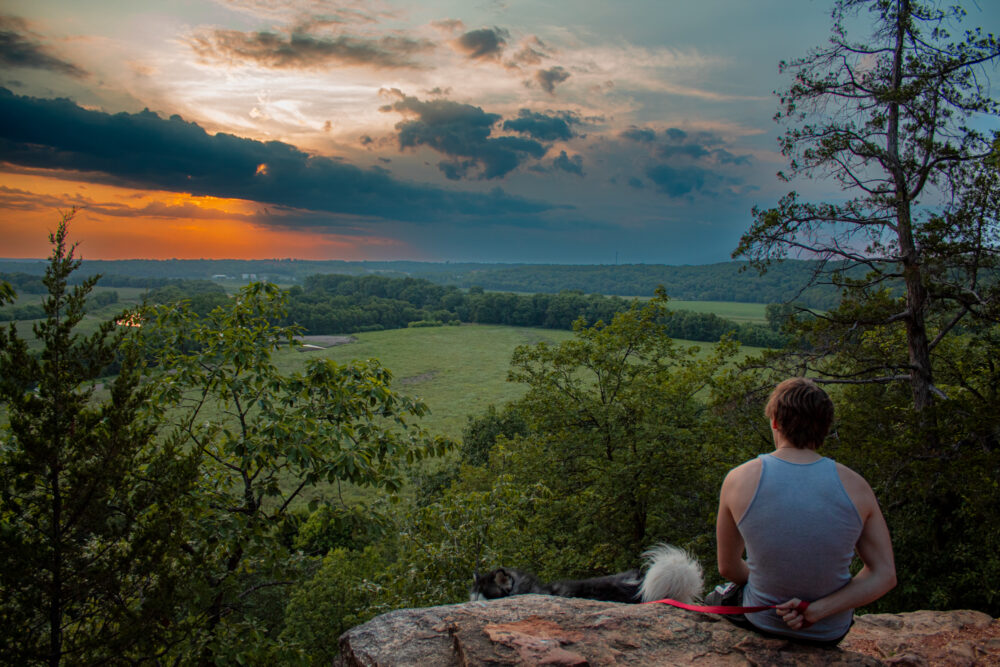 A man sits on a rock with his dog overlooking a sunset with a stormy sky in the distance.