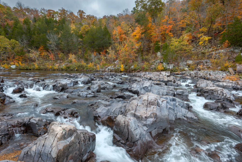 Large chunks of exposed granite rise out of a riverbed. Water rushes around the granite as it flows downstream. In the background, a hilly riverbank rises up, covered in trees with foliage transitioning into the fall season.