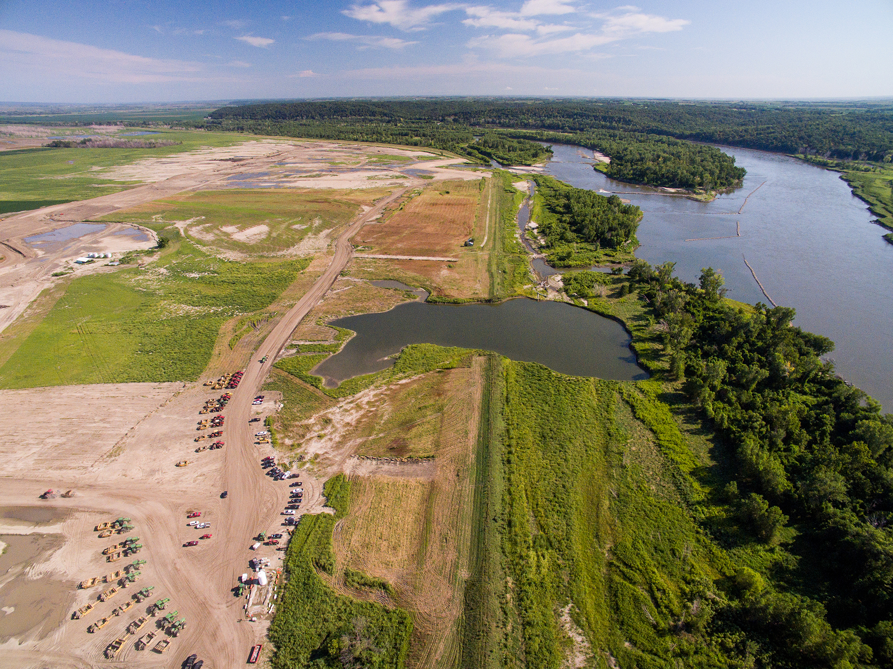 An aerial perspective of a construction site situated along the river levee. The river is on the right. On the left is a large clearing staged with work trucks and construction equipment. The old levee runs up the middle of the image, dividing the water from the land.