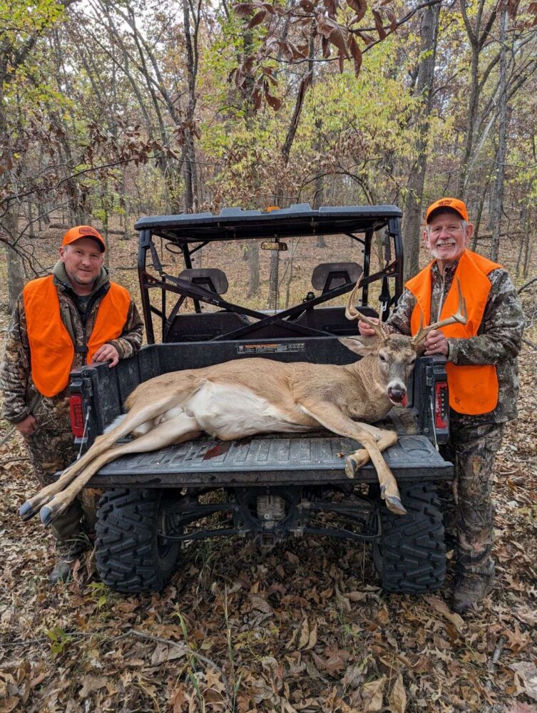 Photo of two men showcasing a buck on the back of a Gator ATV.