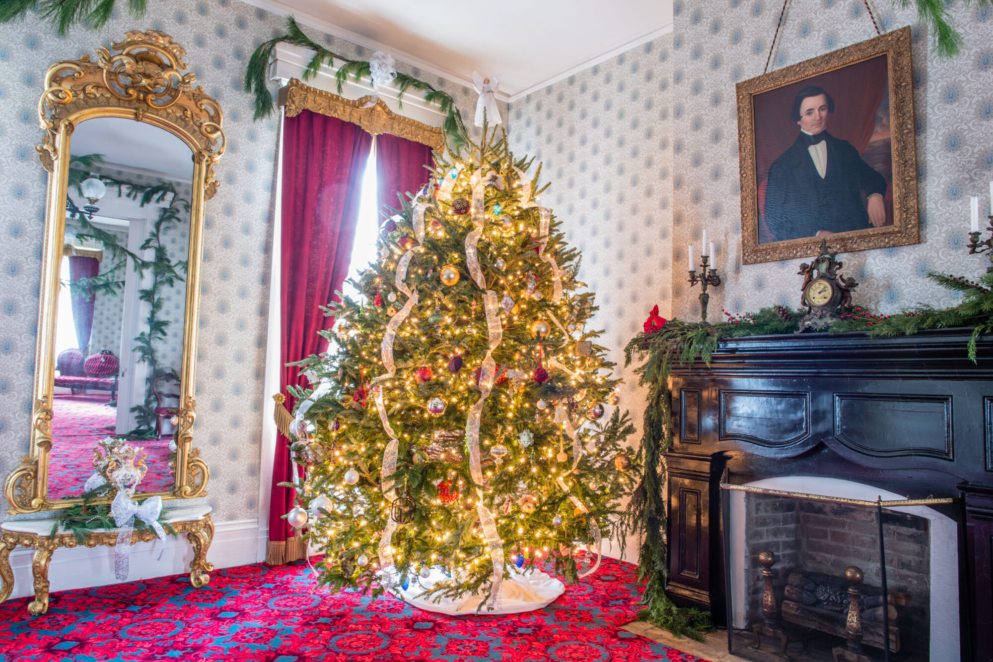 Photo of a large Christmas tree in a corner by a window and fireplace.