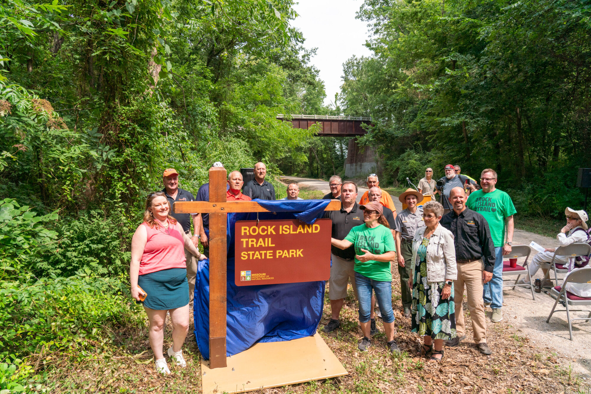 A group of people gather around a sign denoting the Rock Island Trail head, partially covered by a ceremonial sheet.