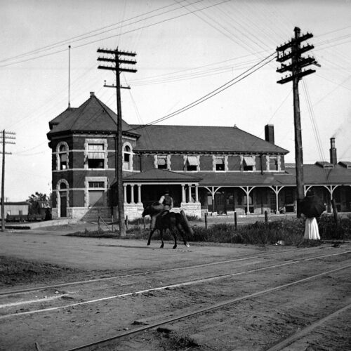 an old photo of a man riding a horse on a road in front of a building over railroad tracks.