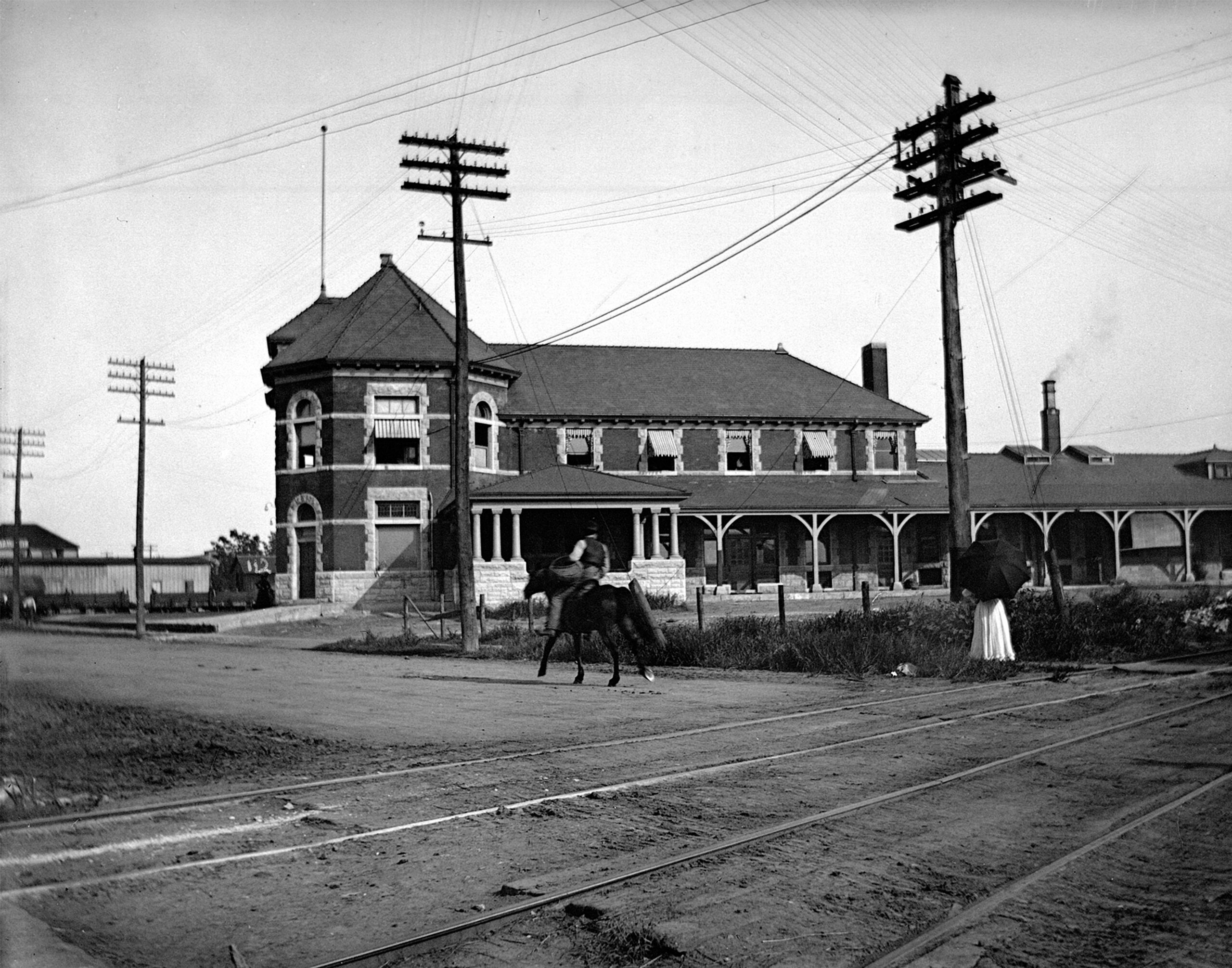 an old photo of a man riding a horse on a road in front of a building over railroad tracks.