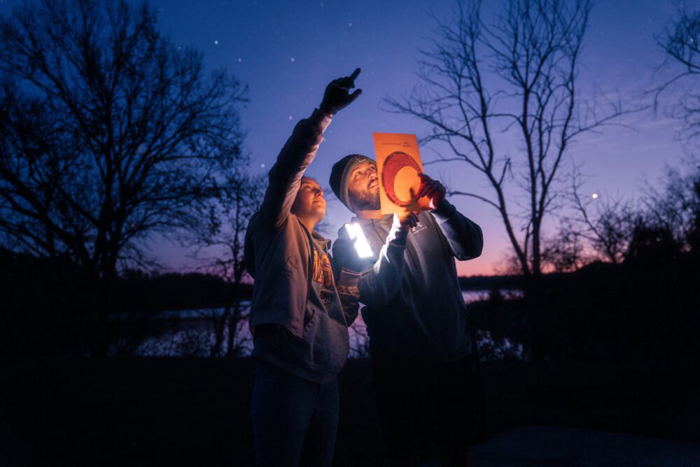 two people look at the night sky while referencing a paper guide.