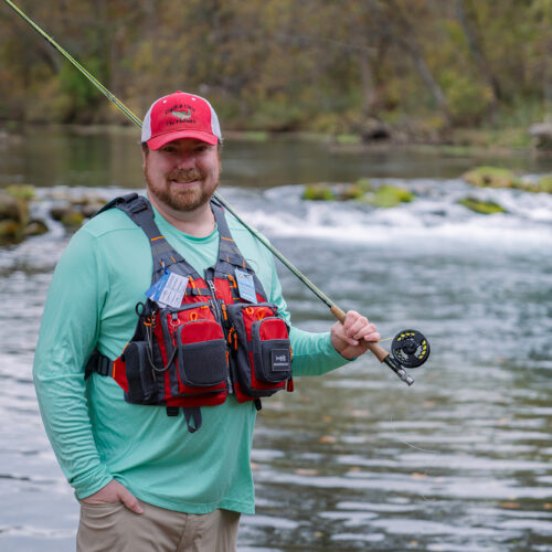 Man with a fishing vest and pole stands in front of a river smiling at the camera