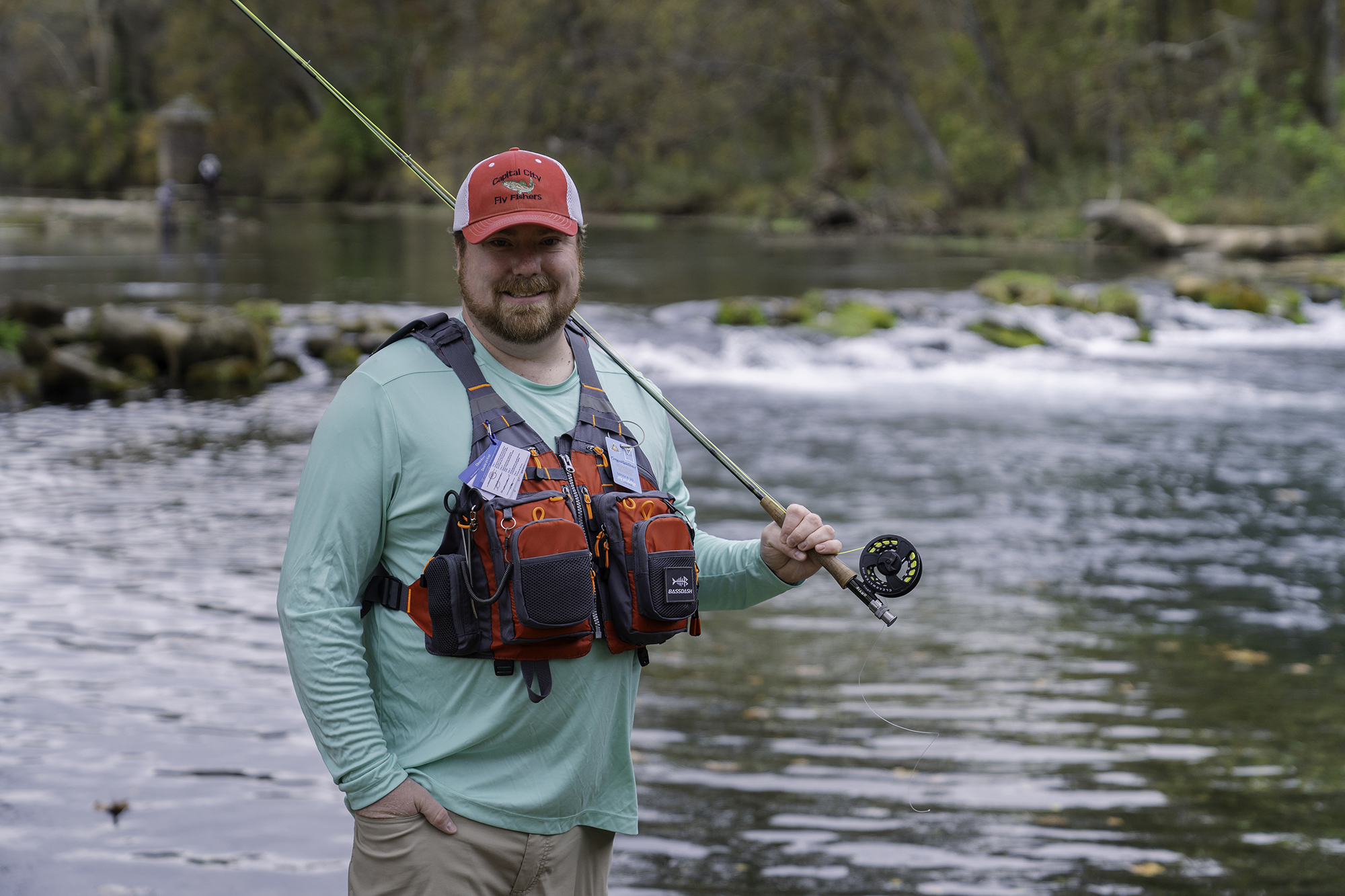 Man with a fishing vest and pole stands in front of a river smiling at the camera