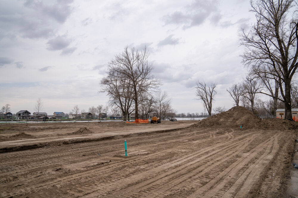 Freshly flattened dirt with tread marks leads to a mound of dirt. Trees and homes are in the distance.