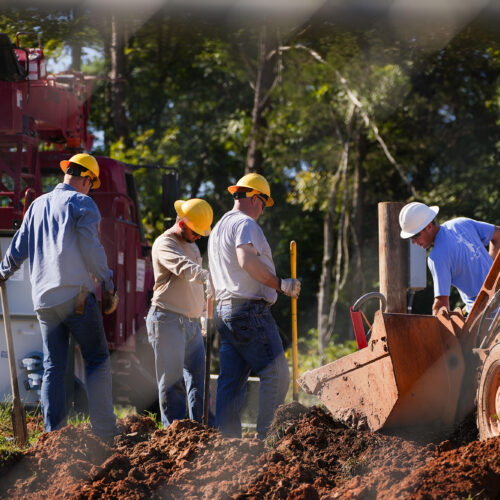 Construction workers gathered around a large back-hoe digging a hole.