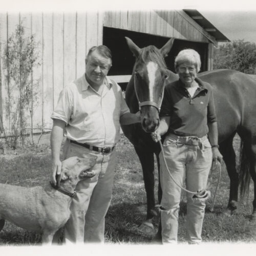 Old photo of a man and woman with a dog and horse, standing in front of a barn.
